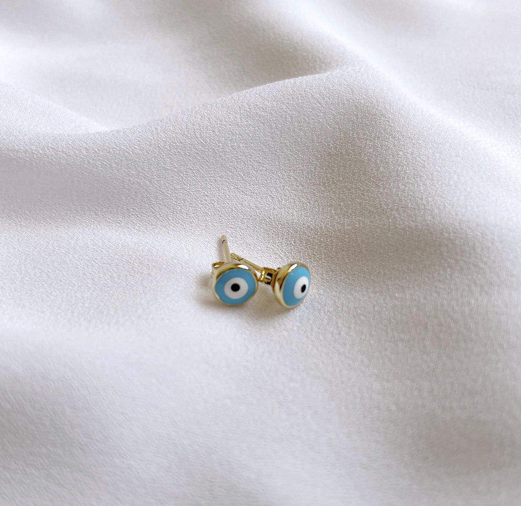 A pair of gold stud earrings with blue and white evil eye designs, resting on a soft, draped white fabric background.