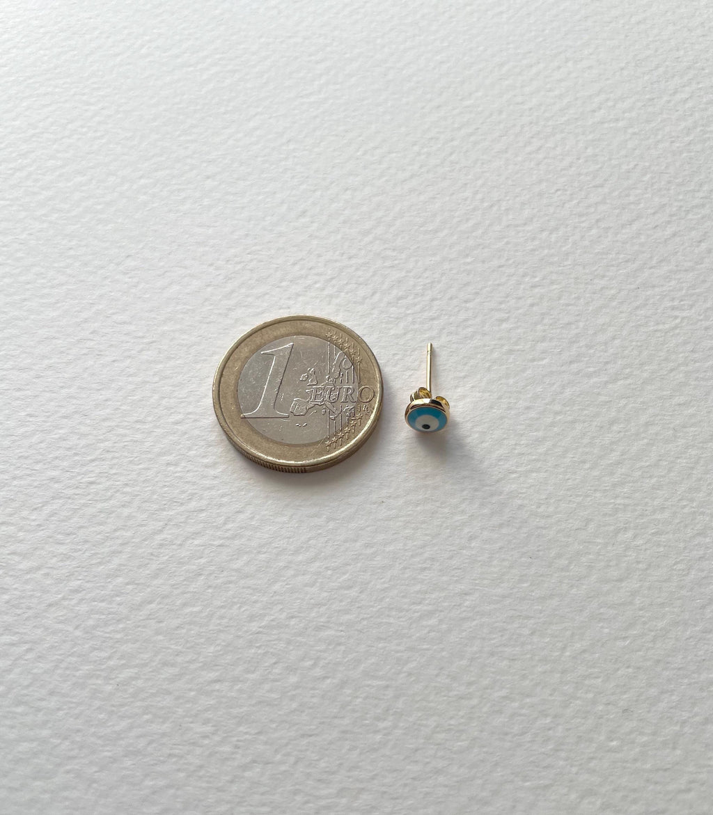 A tiny gold stud earring with a small blue and white evil eye bead, shown next to a 1 Euro coin for scale on a textured white background.