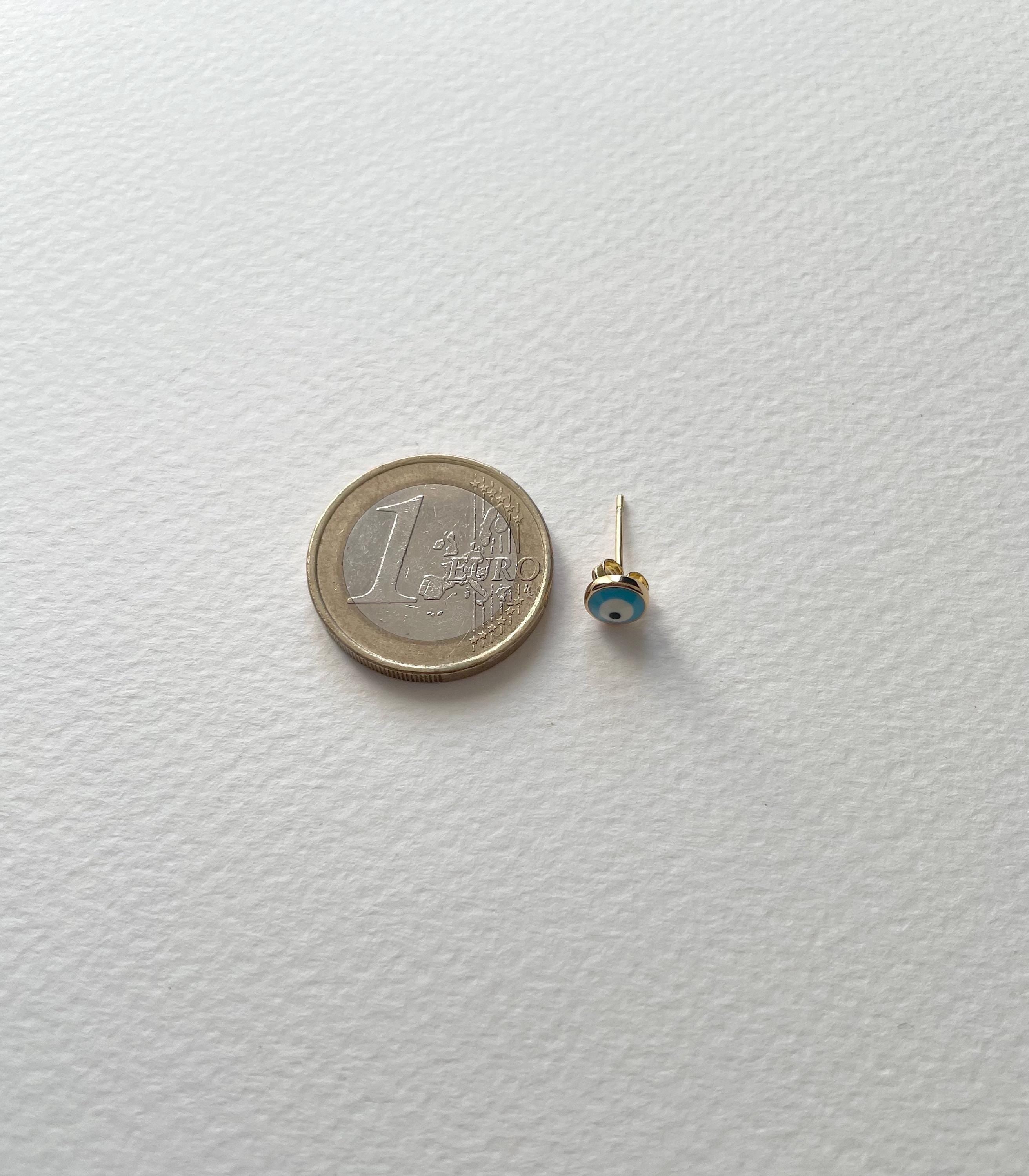 A tiny gold stud earring with a small blue and white evil eye bead, shown next to a 1 Euro coin for scale on a textured white background.