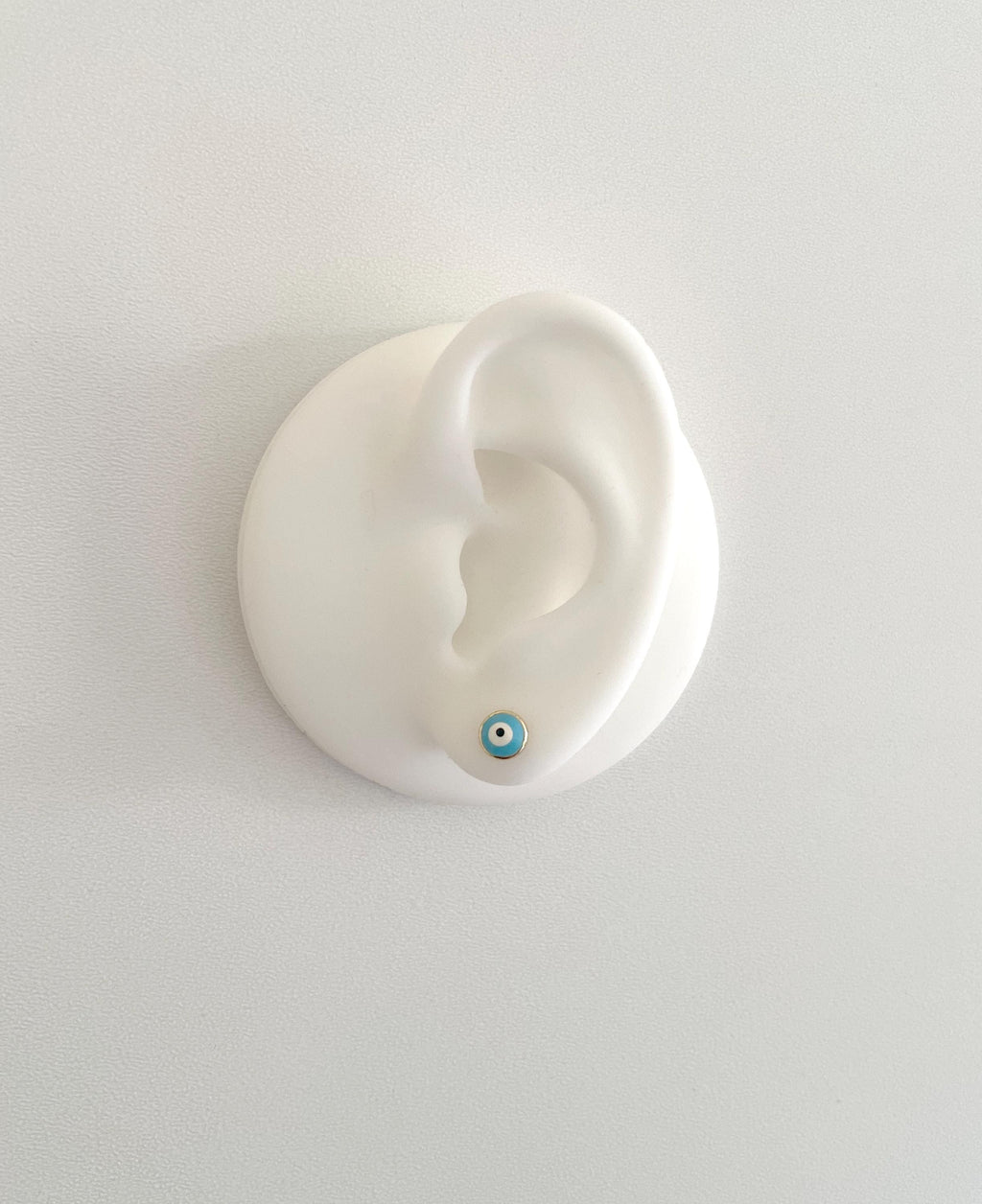 A white silicone ear-shaped wall display holding a small blue and white evil eye stud earring, photographed against a plain light gray wall.