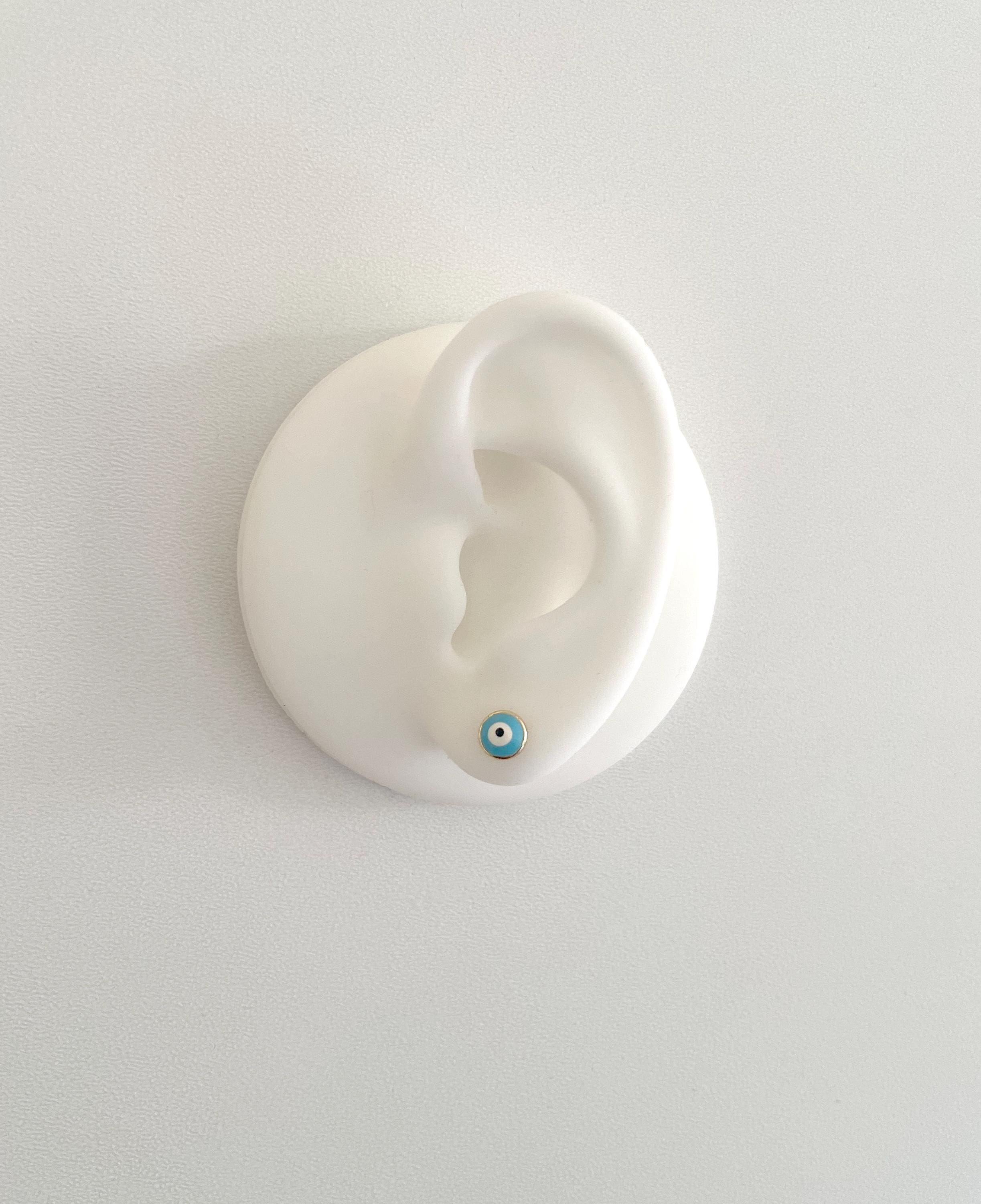 A white silicone ear-shaped wall display holding a small blue and white evil eye stud earring, photographed against a plain light gray wall.