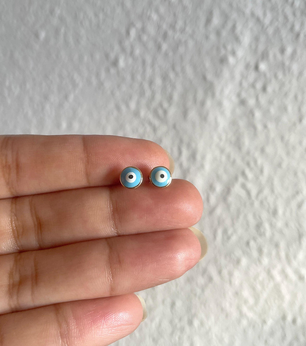 A pair of small blue and white evil eye stud earrings held between a person's fingers against a textured white wall background.