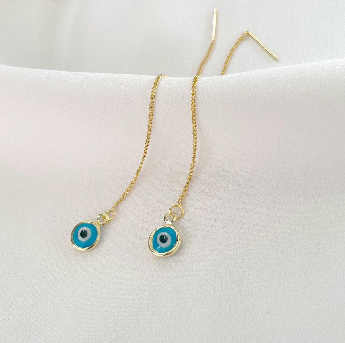 A pair of gold chain earrings with circular evil eye pendants, displayed on a white fabric surface.