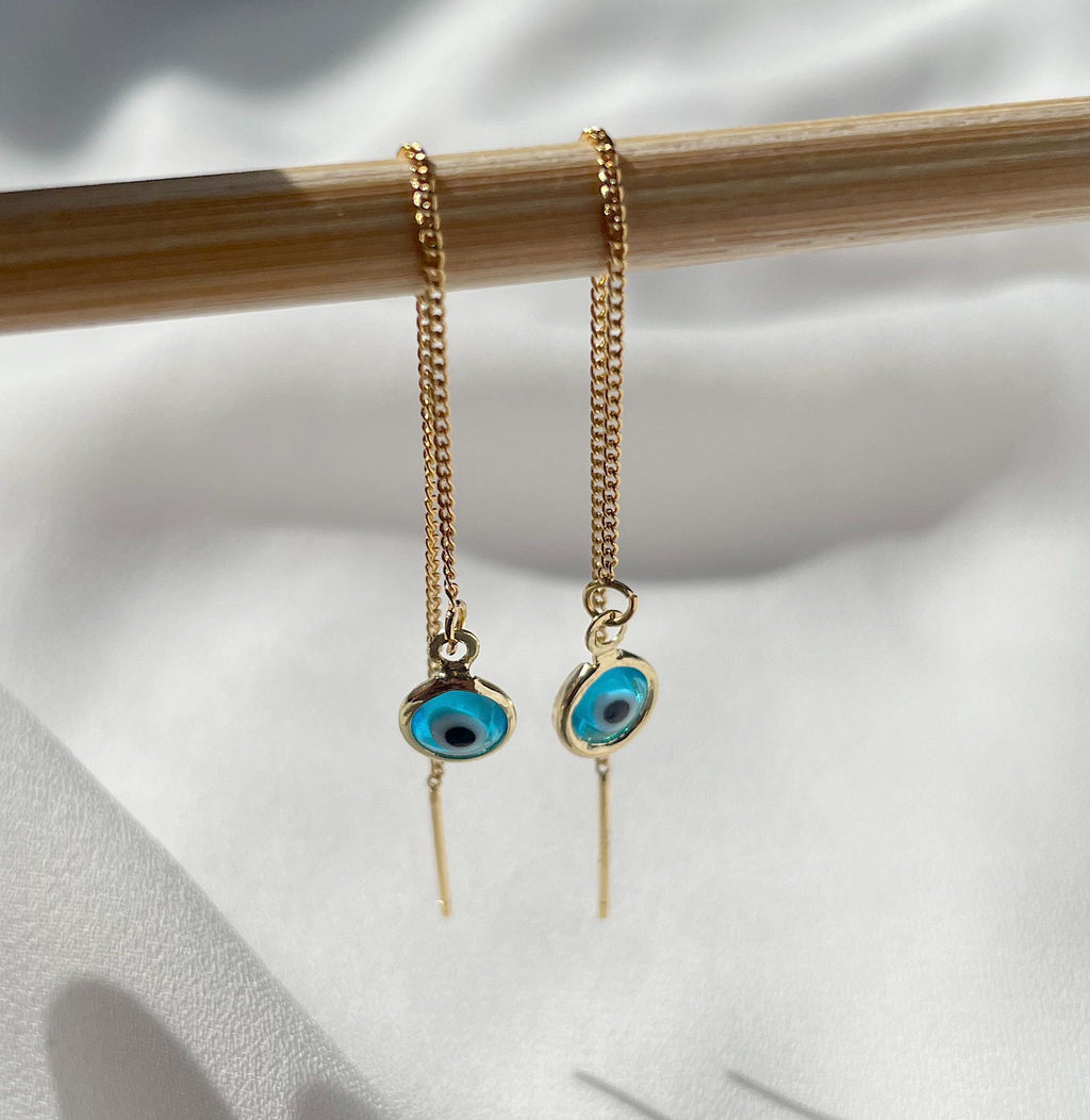 A pair of gold chain drop earrings with blue and black evil eye charms, hanging from a wooden rod against a white textured fabric background.