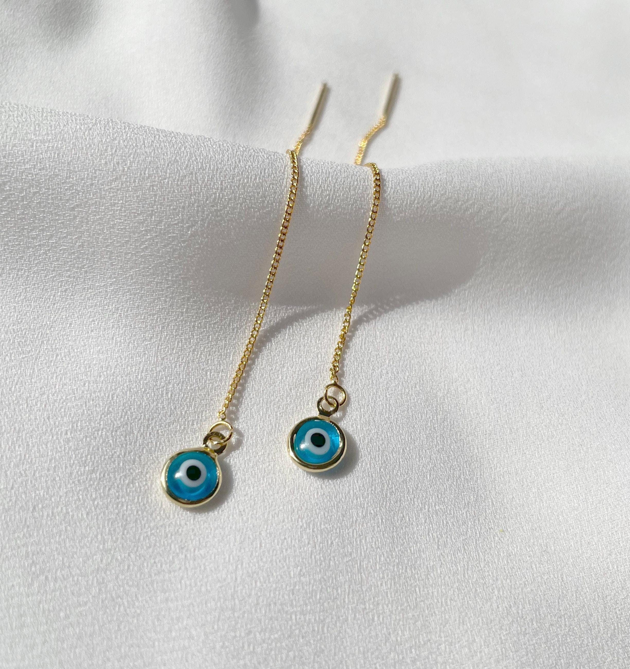 A pair of gold chain drop earrings with round blue evil eye charms, displayed on a soft white fabric background.