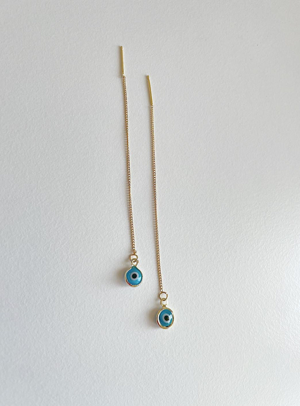 A pair of long gold chain earrings with small evil eye charms, displayed against a plain light gray background.