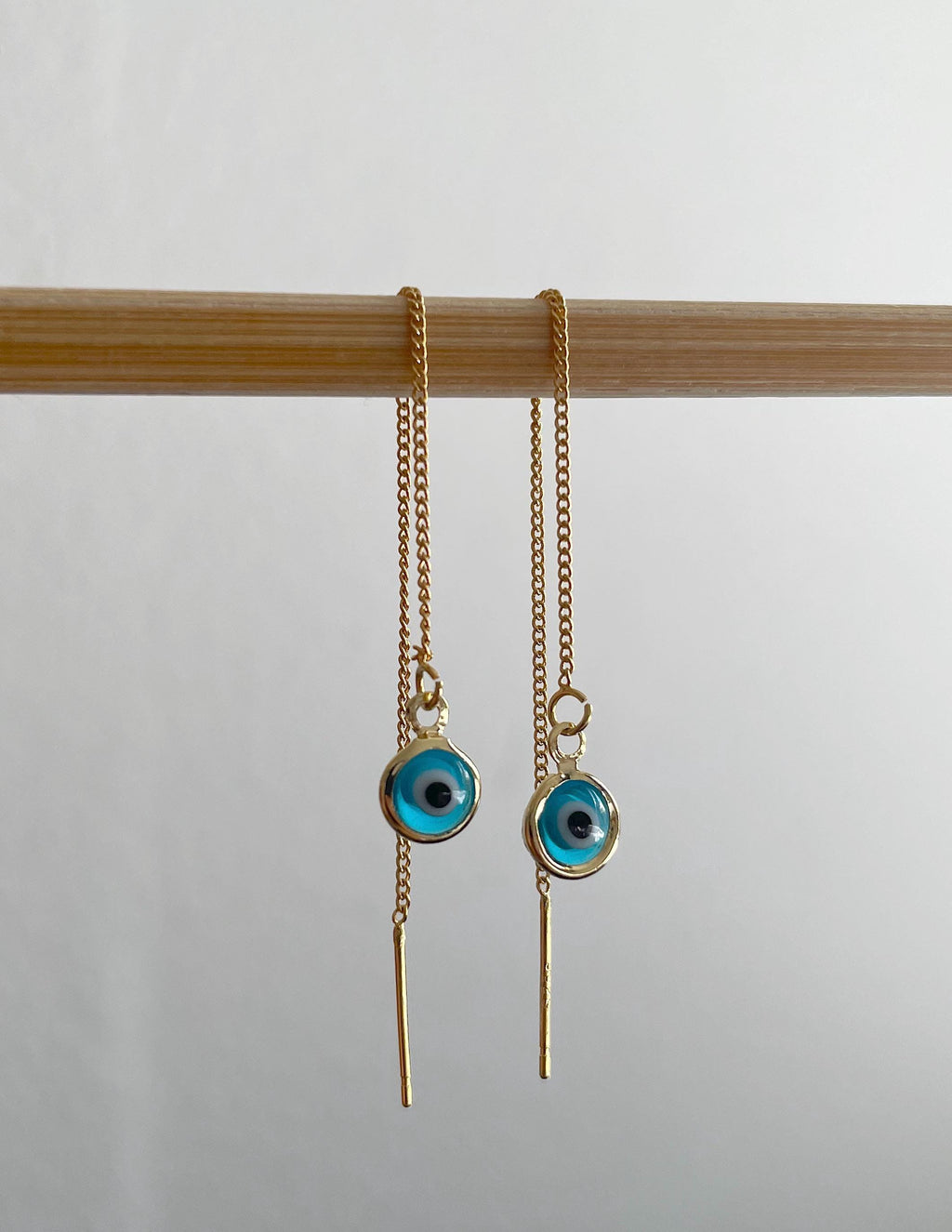A pair of gold-toned dangle earrings with circular turquoise blue evil eye charms hanging from a light wood rod against a plain light gray background.