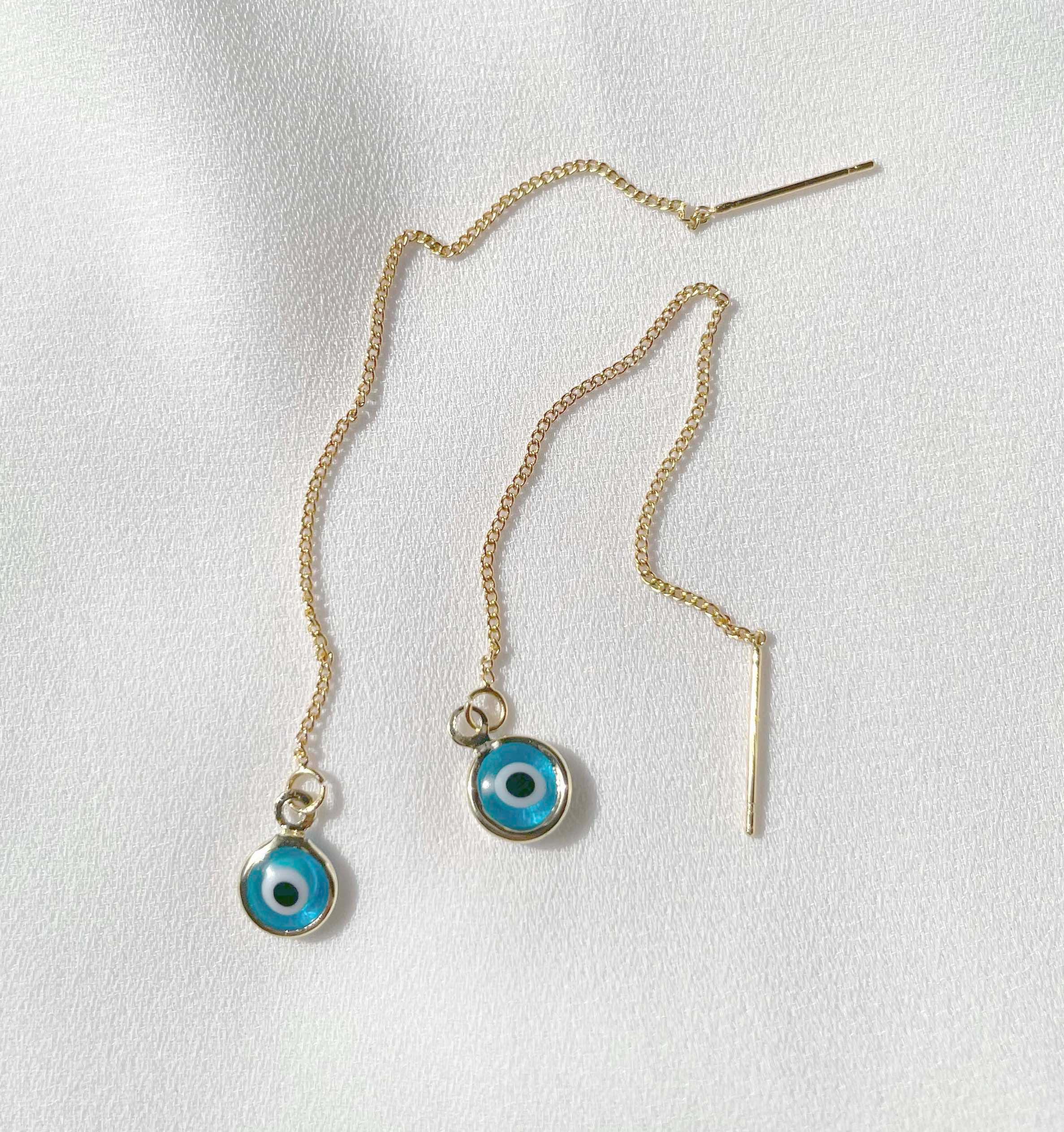 A pair of gold-toned chain earrings with round evil eye charms featuring blue, white, and black details, displayed on a textured white fabric background.