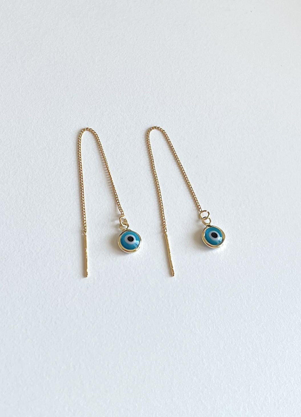 A pair of gold-toned drop earrings with long chains and small circular evil eye charms featuring blue and black details, displayed on a light off-white background.