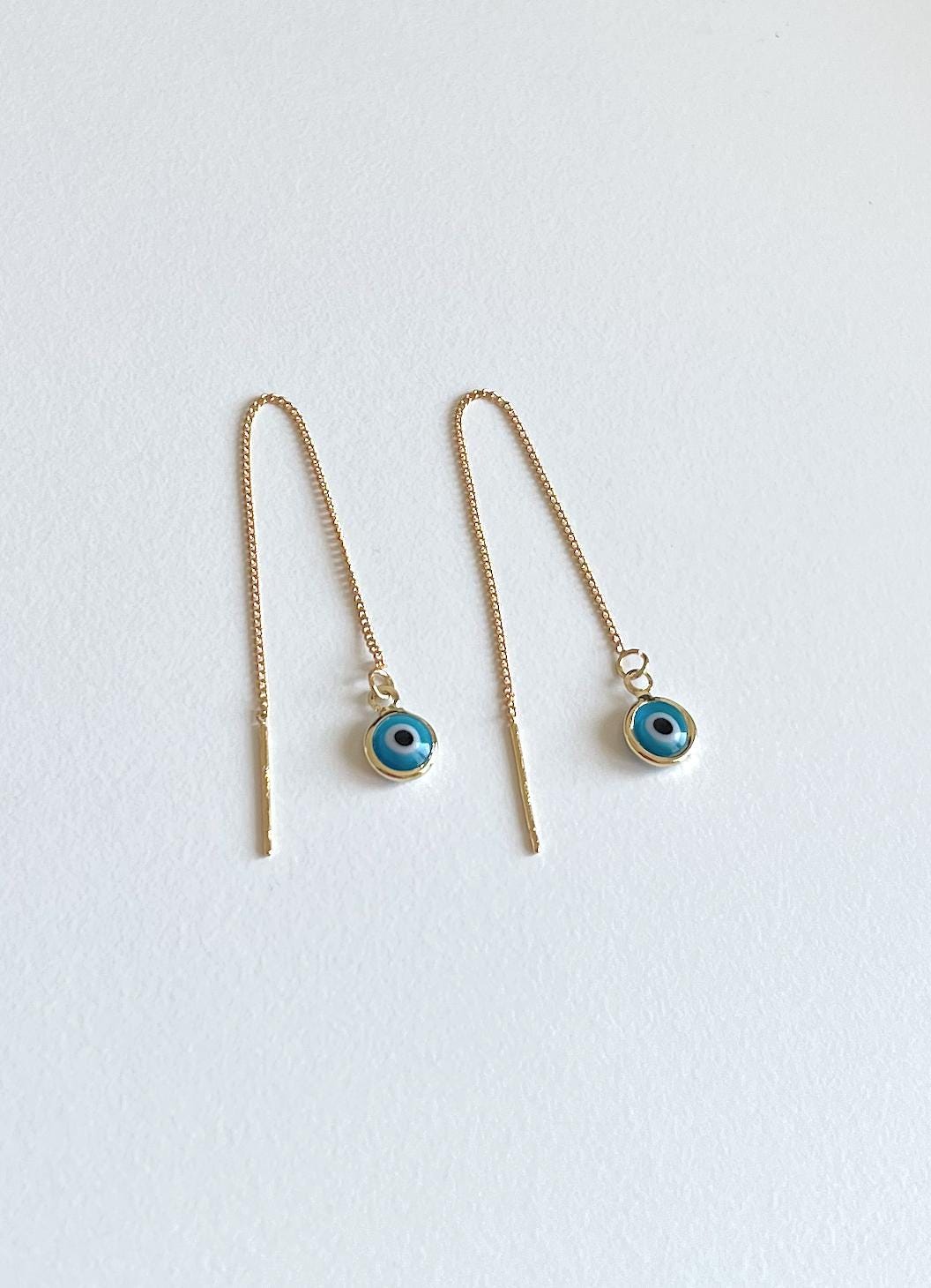A pair of gold-toned drop earrings with long chains and small circular evil eye charms featuring blue and black details, displayed on a light off-white background.