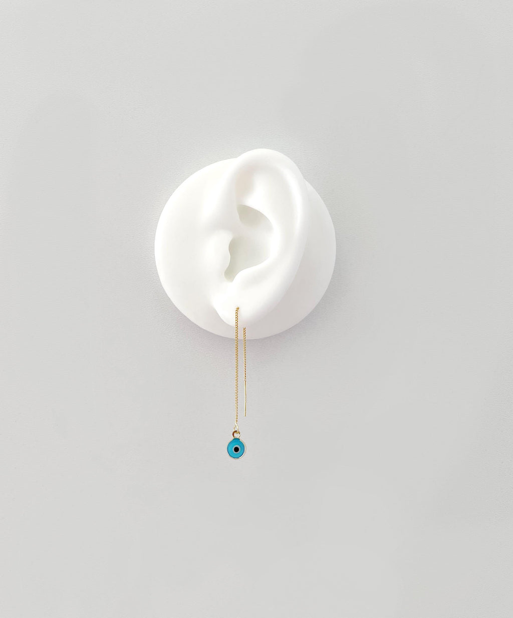 A delicate gold dangle earring with a small turquoise blue evil eye bead, displayed on a white ear-shaped stand against a light gray background.