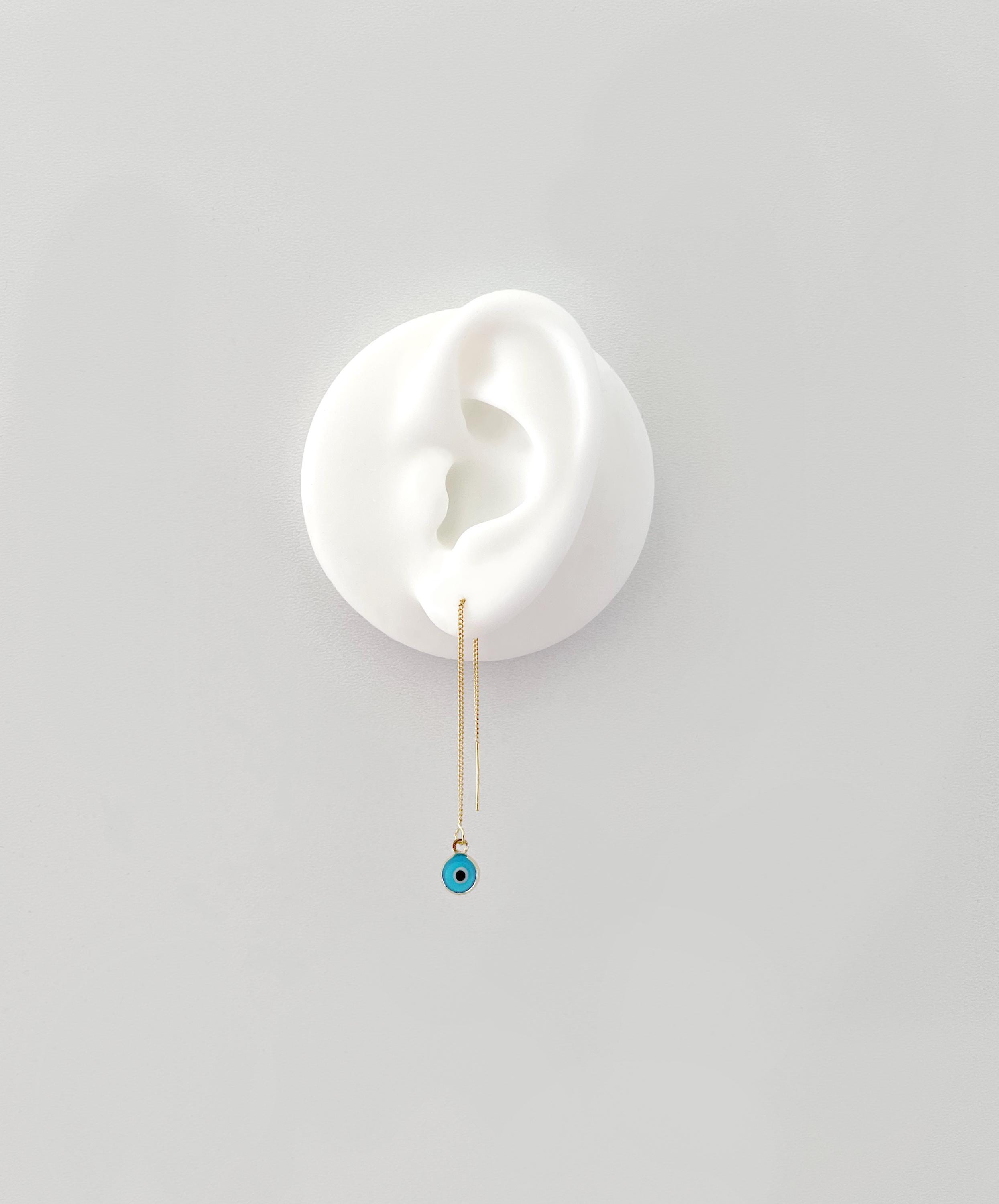 A delicate gold dangle earring with a small turquoise blue evil eye bead, displayed on a white ear-shaped stand against a light gray background.