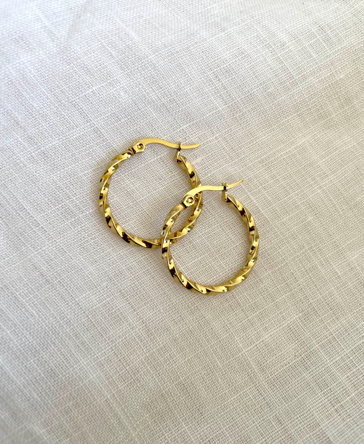 A pair of textured gold hoop earrings with fishhook backs, displayed on a light beige linen fabric background.