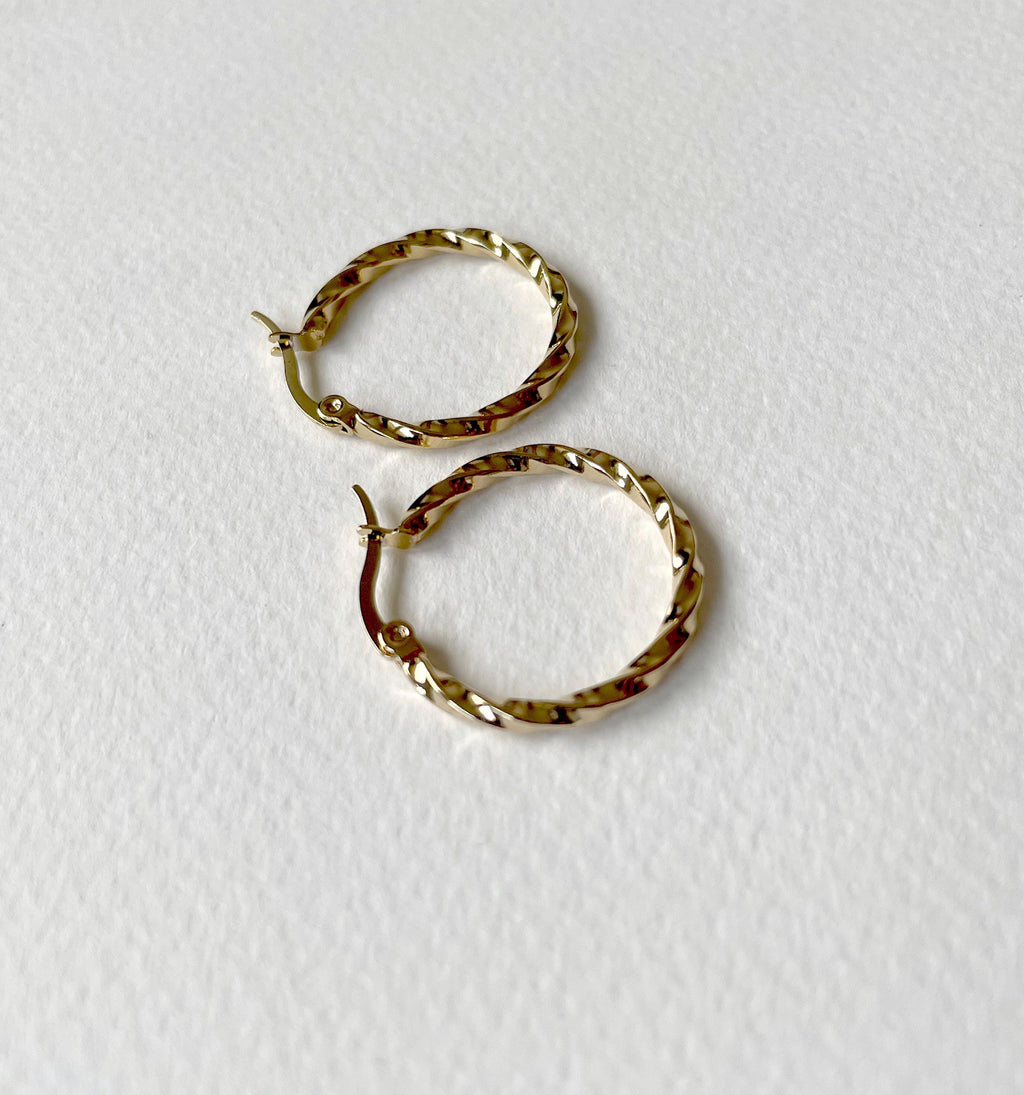 A pair of small gold hoop earrings with a textured, twisted surface, displayed on a textured white background.