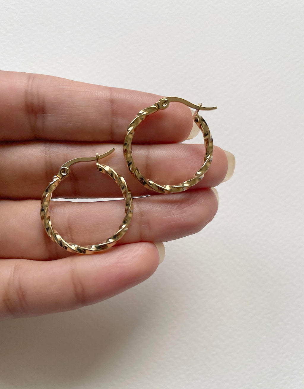 A pair of gold hoop earrings with a textured, woven-like pattern, held in a person's hand against a plain off-white background.
