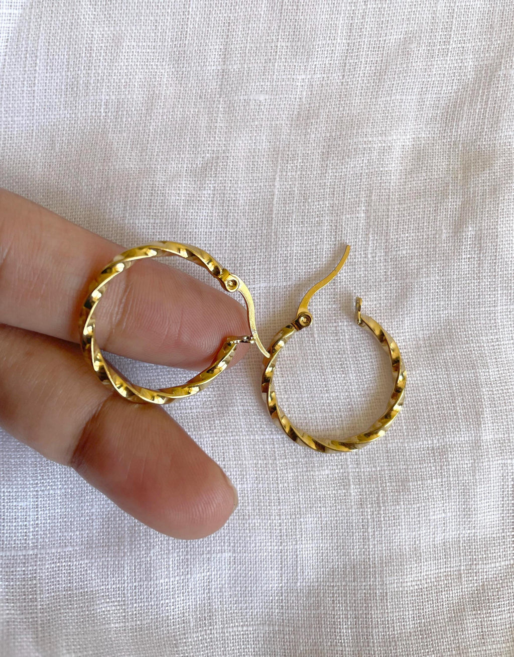 A pair of textured gold hoop earrings held between fingers against a white fabric background.