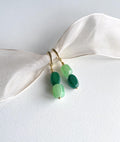 A pair of gold-tone drop earrings with three green beads of varying shades, displayed on a white textured surface with a cream satin ribbon.