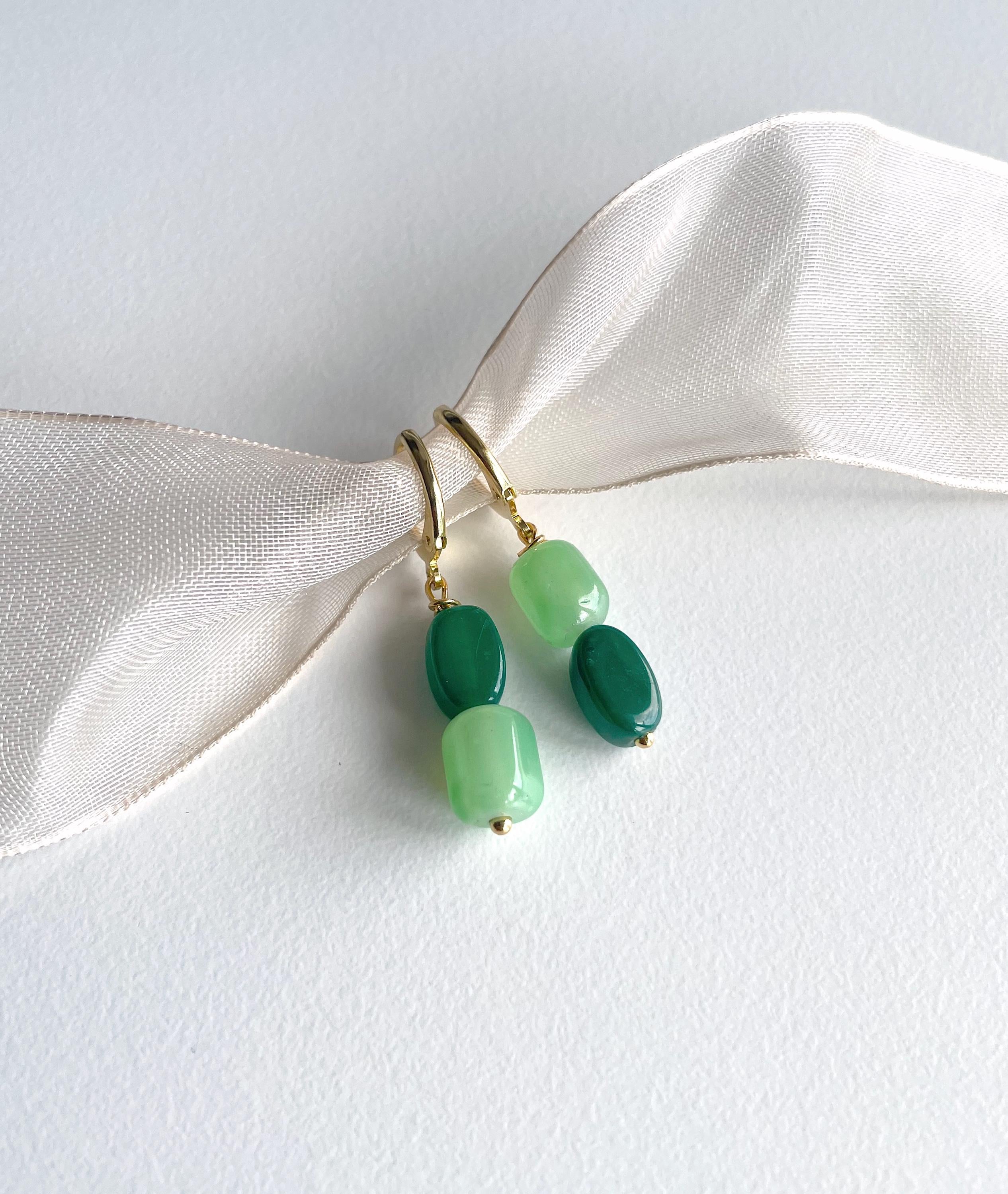 A pair of gold-tone drop earrings with three green beads of varying shades, displayed on a white textured surface with a cream satin ribbon.