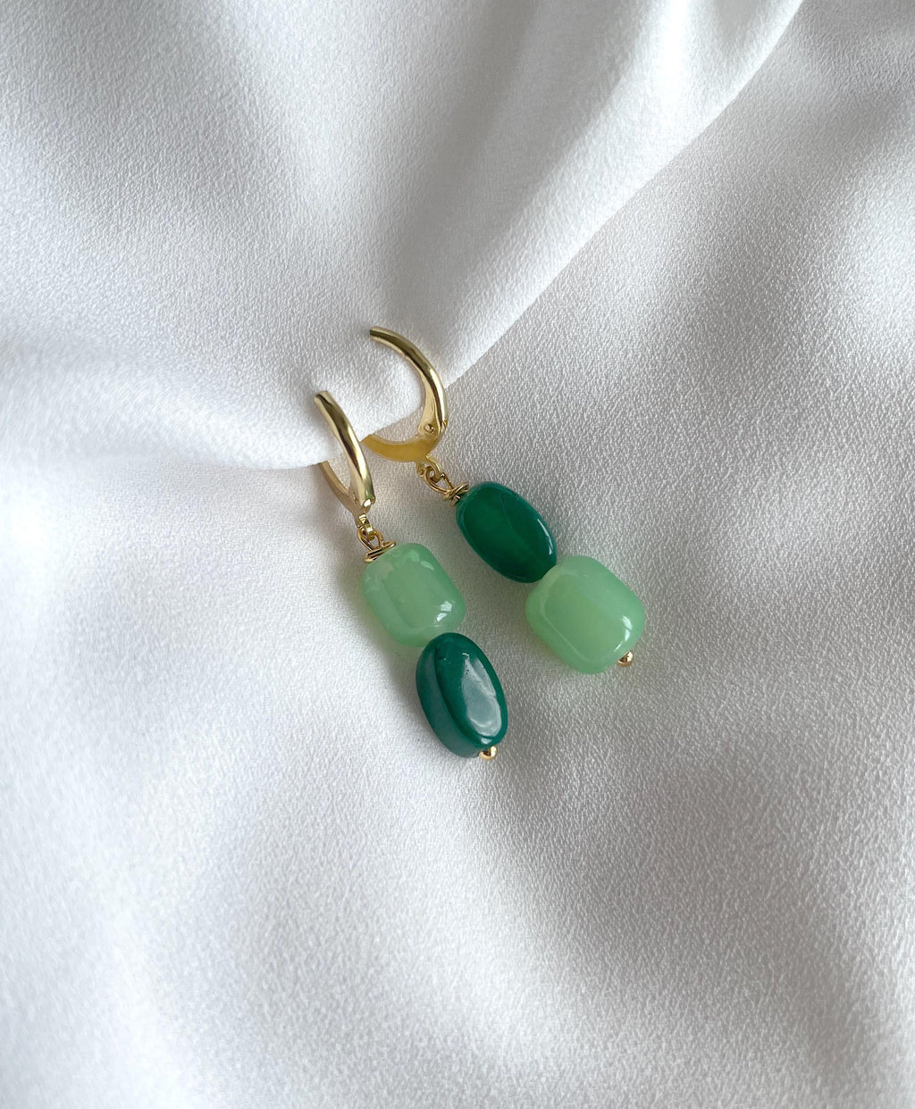 A pair of gold hoop earrings with green beads, displayed on a draped white fabric background.
