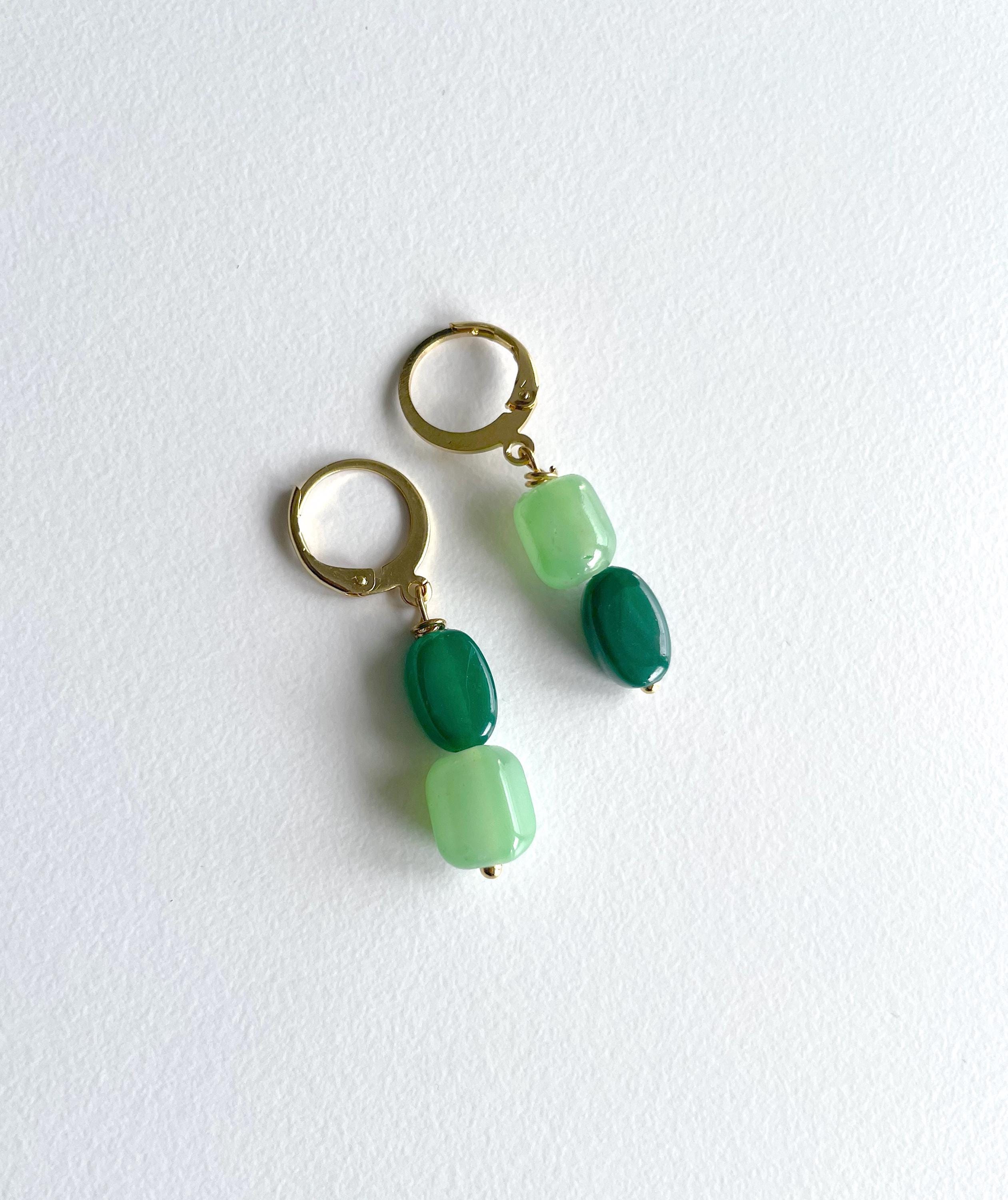 A pair of gold hoop earrings with dangling green beads, featuring one dark green oval bead and one light green rounded rectangular bead, displayed on a textured white background.