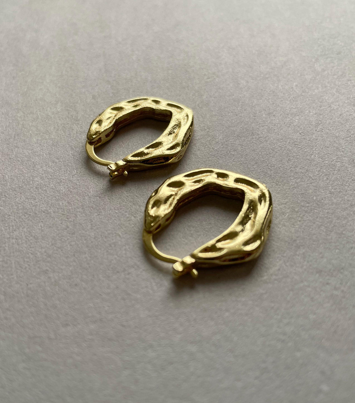 A pair of textured gold plated brass huggie hoop earrings with organic, wavy edges, displayed on a neutral gray surface.