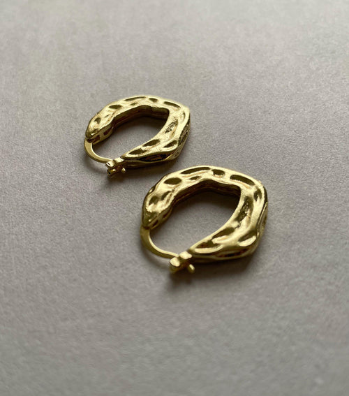 A pair of textured gold plated brass huggie hoop earrings with organic, wavy edges, displayed on a neutral gray surface.