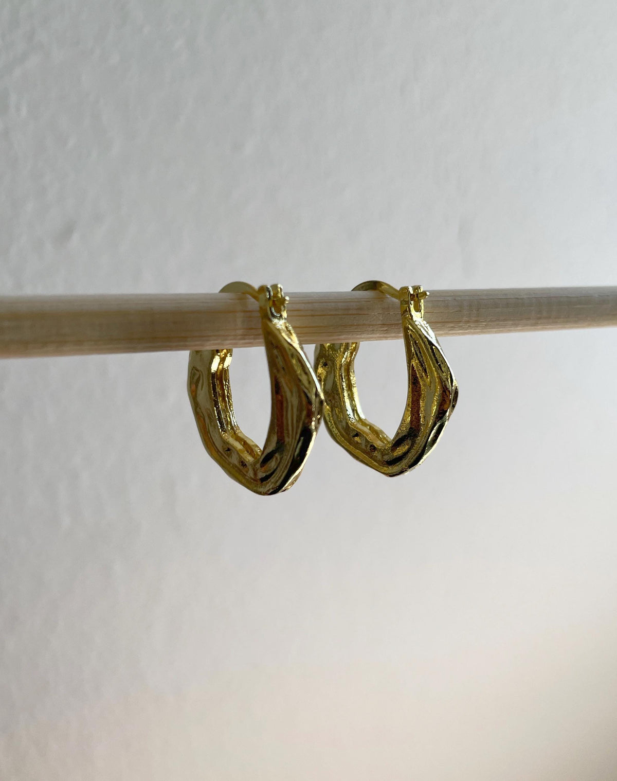 A pair of hammered gold plated brass hoop earrings hanging from a light-colored wooden rod against a plain off-white wall.