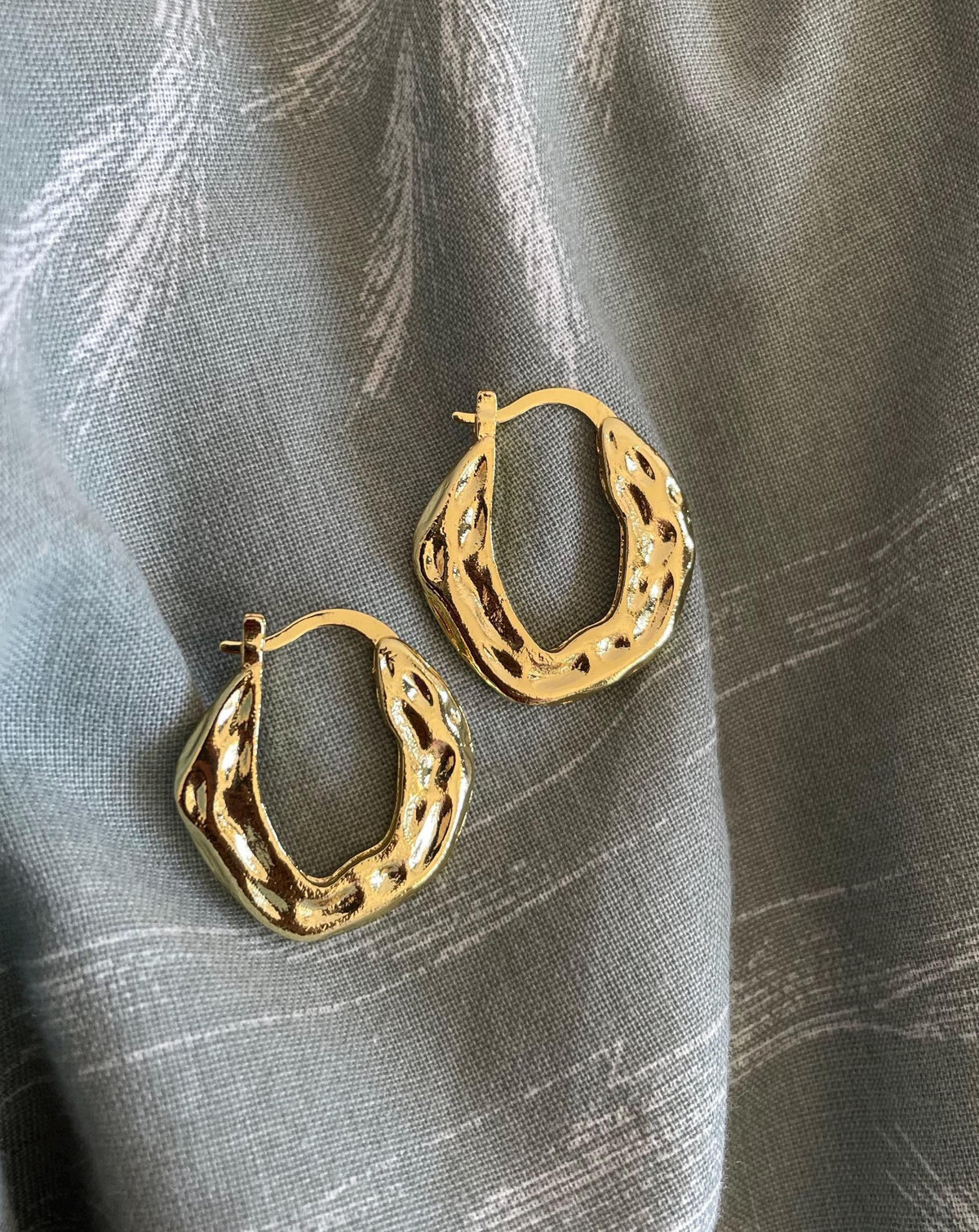 A pair of textured gold hoop earrings with organic, wavy contours and openwork detailing, displayed on a draped gray fabric background.