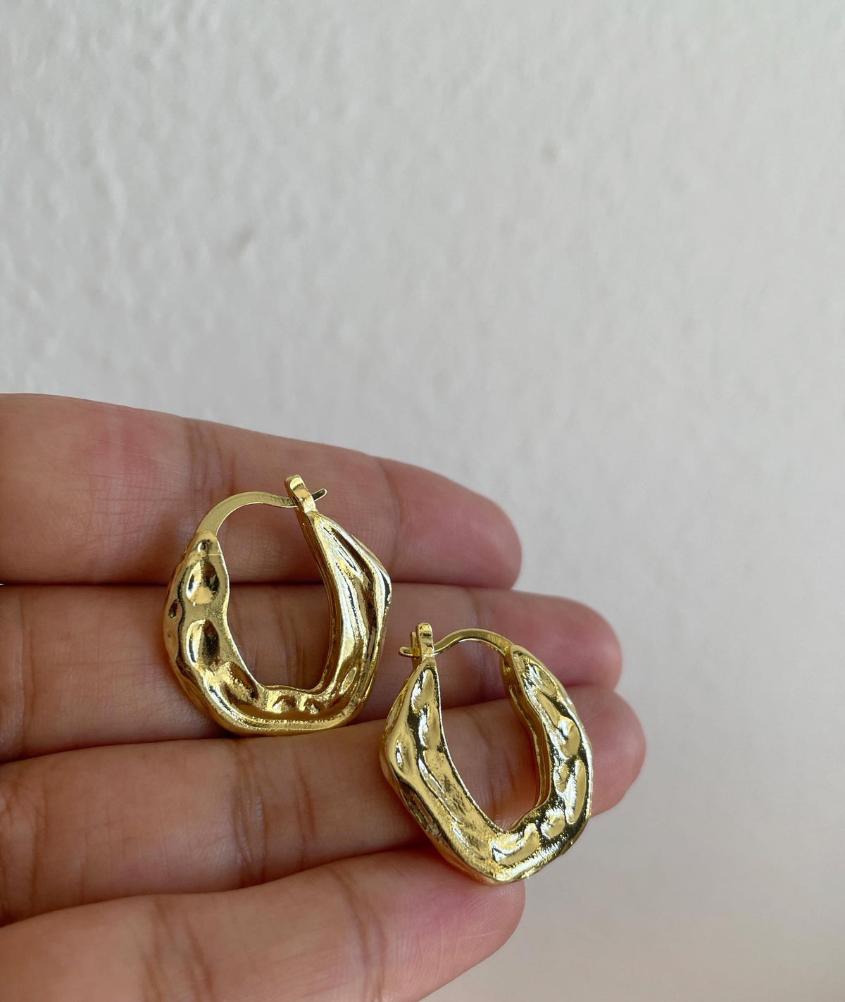 A pair of textured hammered gold plated brass hoop earrings held in a person's hand against a plain off-white wall background.