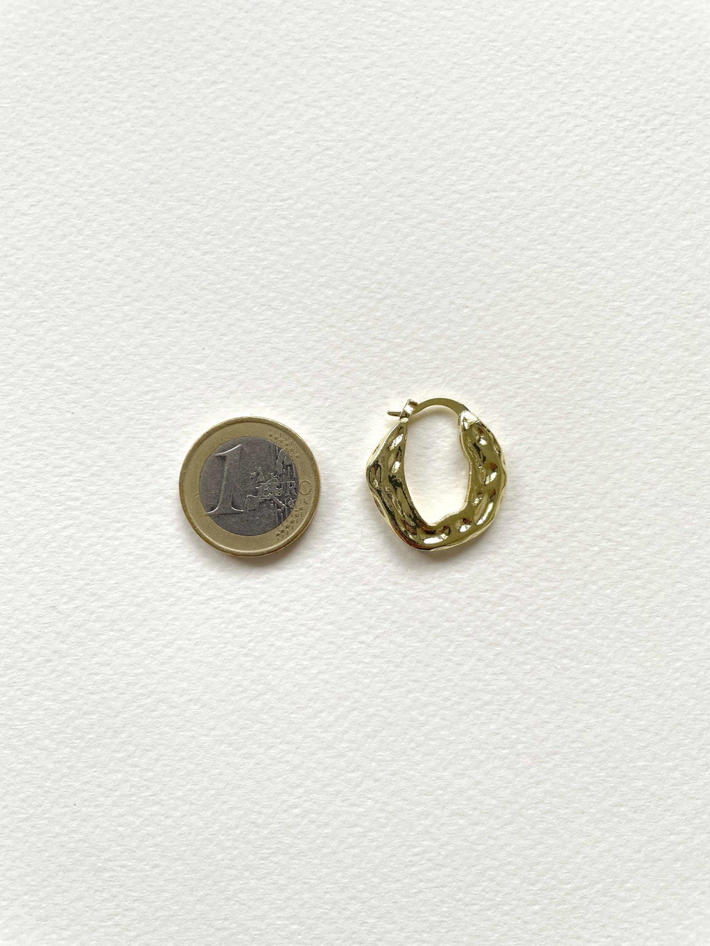 A small gold plated huggie hoop earring shown next to a 1 Euro coin for scale, on a textured white background.