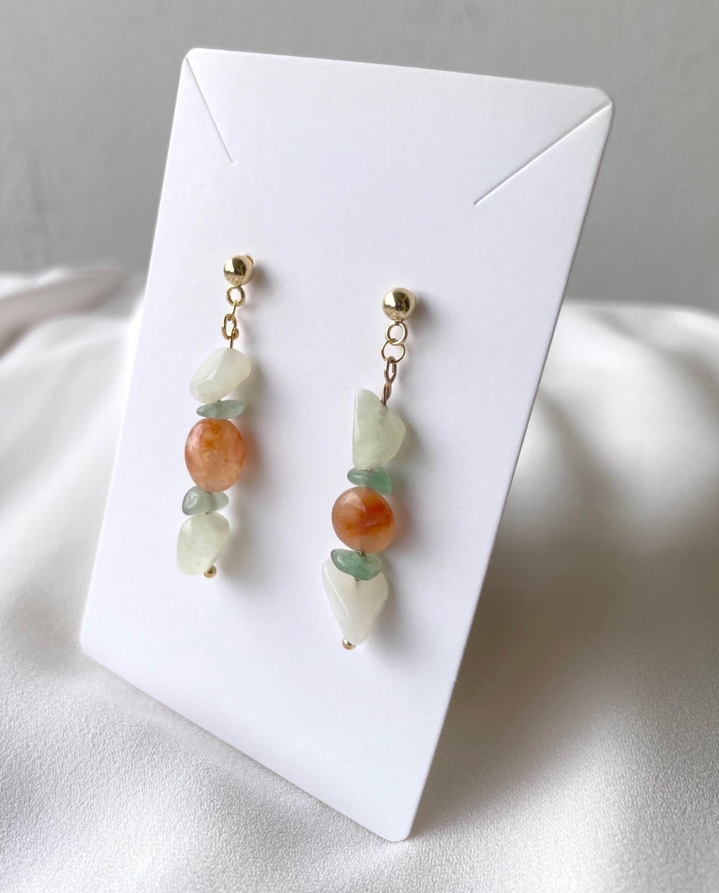 A pair of handmade dangle earrings with gold-toned posts, featuring a combination of light green jade beads, a larger peach-orange stone bead, and white stone beads, displayed on a white card.