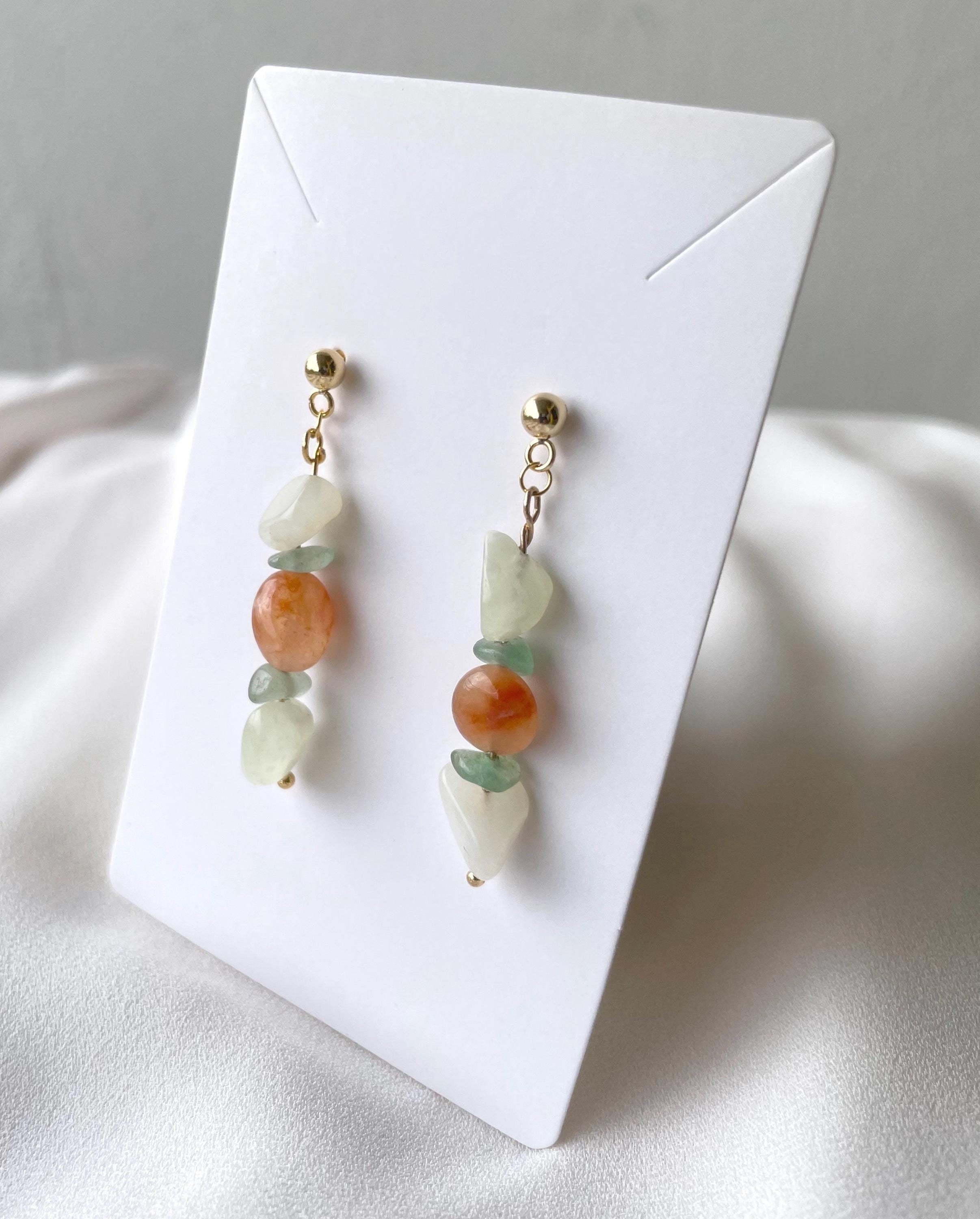 A pair of handmade dangle earrings with gold-toned posts, featuring a combination of light green jade beads, a larger peach-orange stone bead, and white stone beads, displayed on a white card.
