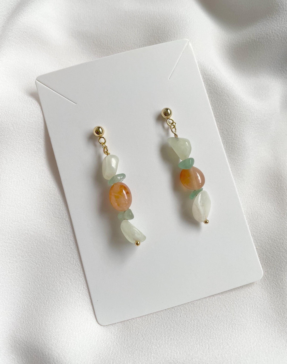 A pair of dangling earrings displayed on a white card against a soft white fabric background. Each earring has gold plated metal with white, pale green, and peach-orange natural gemstone beads.