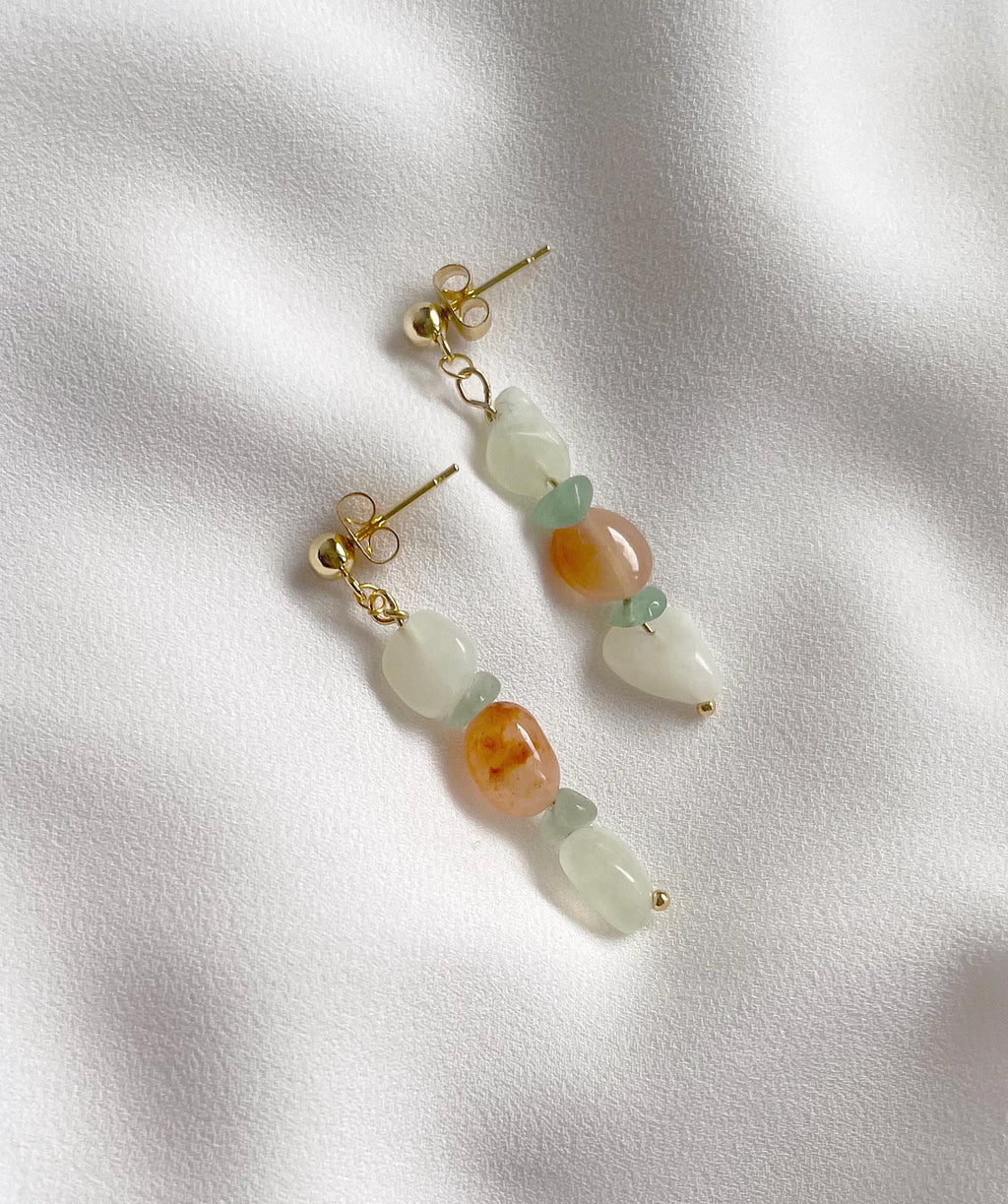 A pair of gold-tone dangle earrings with organic-shaped beads in pale green, peach-orange, and translucent white, displayed on a textured white fabric background.