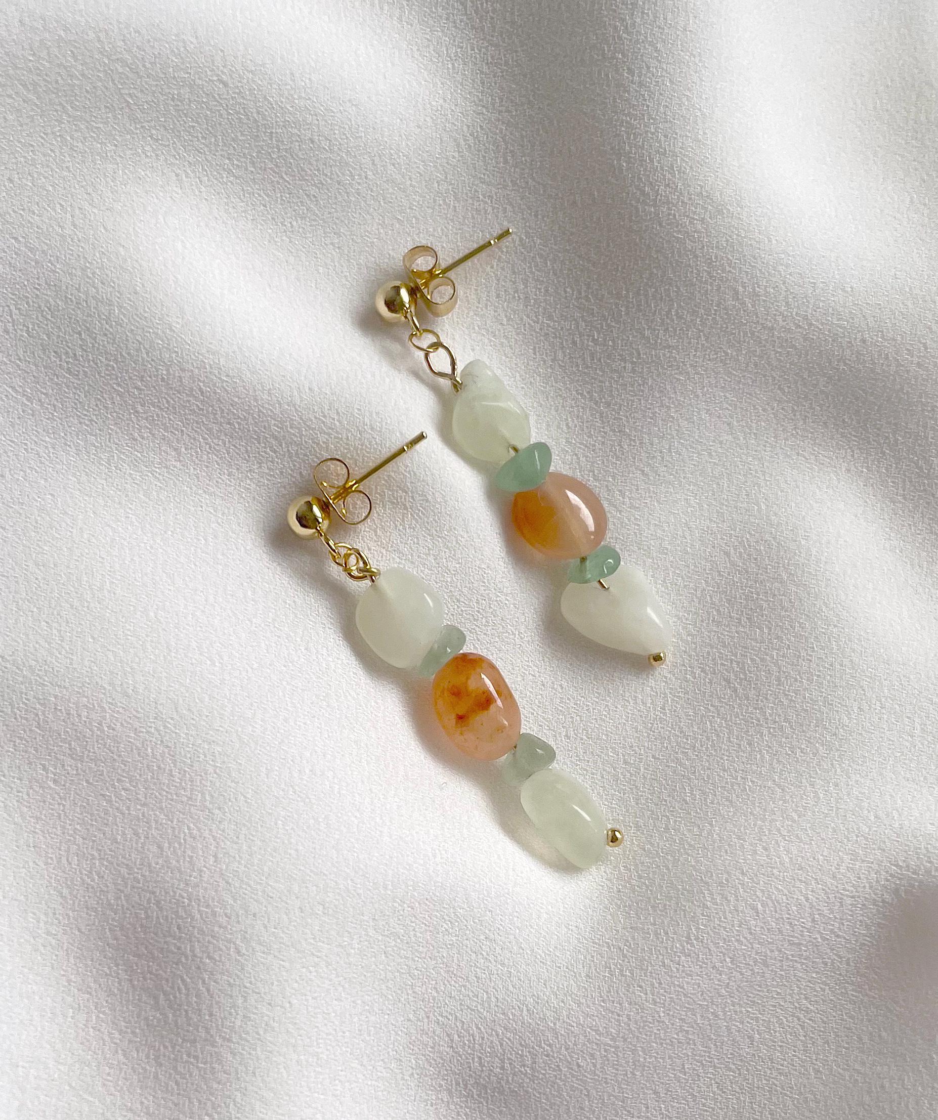 A pair of gold-tone dangle earrings with organic-shaped beads in pale green, peach-orange, and translucent white, displayed on a textured white fabric background.