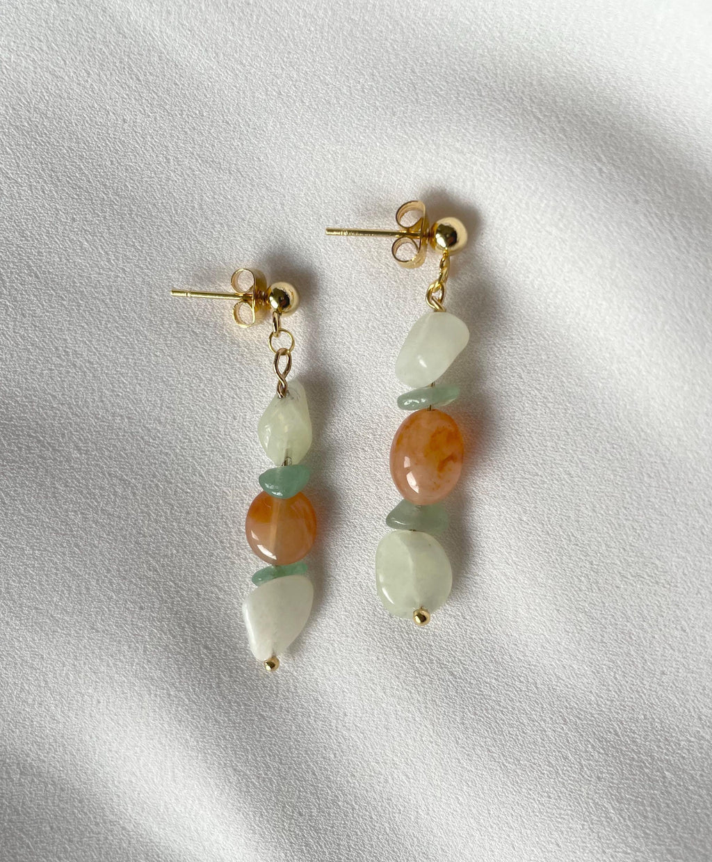 A pair of gold drop earrings with polished stones, featuring light green jade beads, peach-orange agate-like beads, and off-white teardrop beads, displayed on a textured white fabric background.