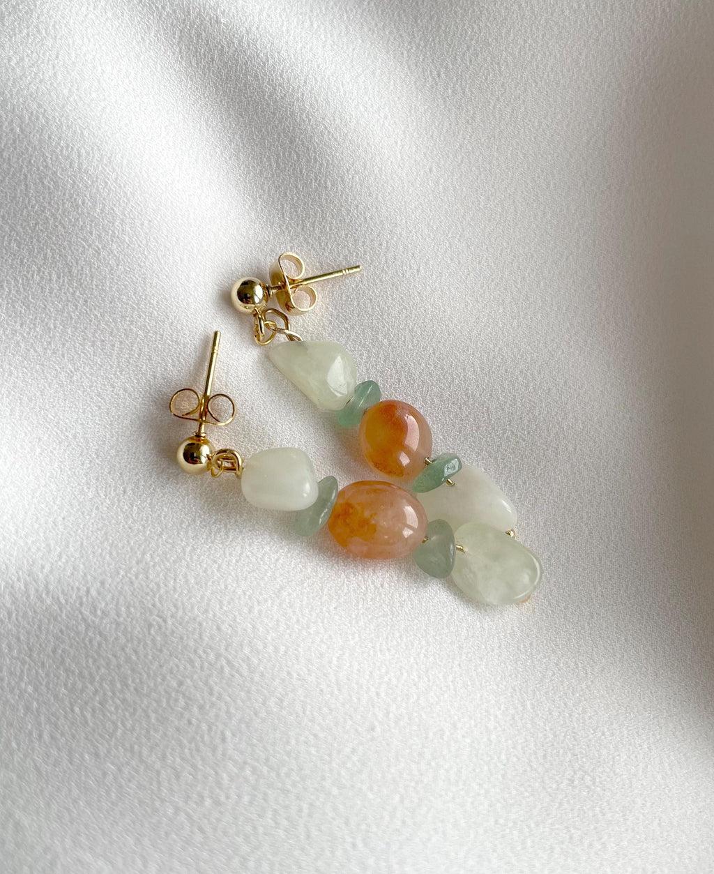 A pair of gold-tone dangle earrings with organic-shaped beads in pale green, translucent white, and peach-orange, displayed on a textured white fabric background.