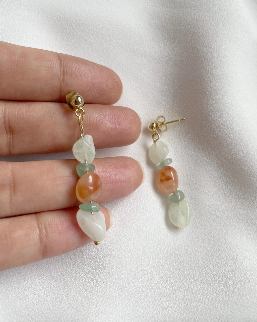 A pair of gold-tone drop earrings with organic-shaped beads in white, light green, and peach-orange, held by a hand against a soft white fabric background.