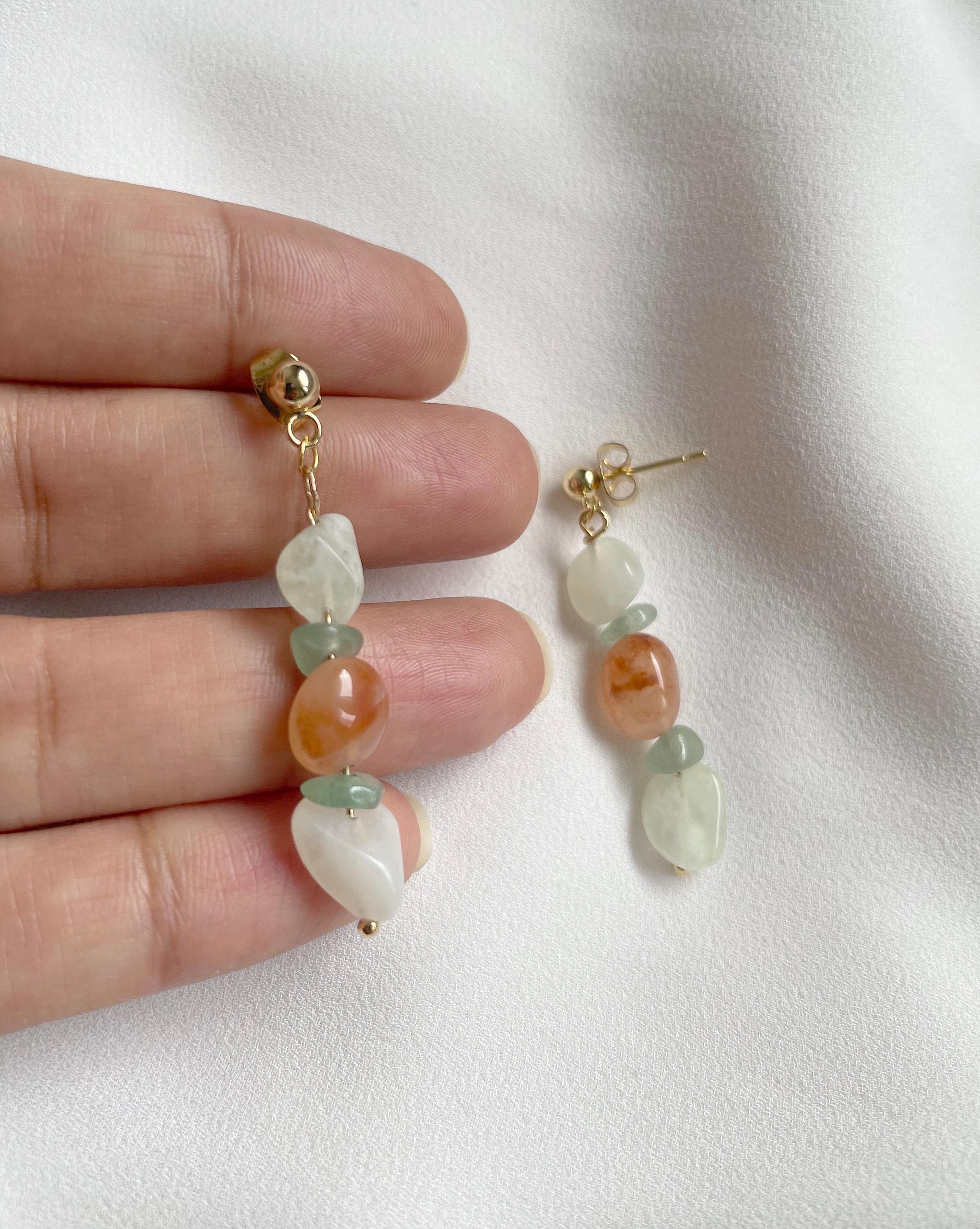 A pair of gold-tone drop earrings with organic-shaped beads in white, light green, and peach-orange, held by a hand against a soft white fabric background.