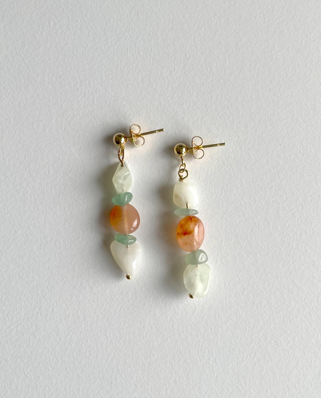 A pair of gold-tone dangle earrings with irregular white beads, light green beads, and round orange stone beads, displayed on a textured light gray background.