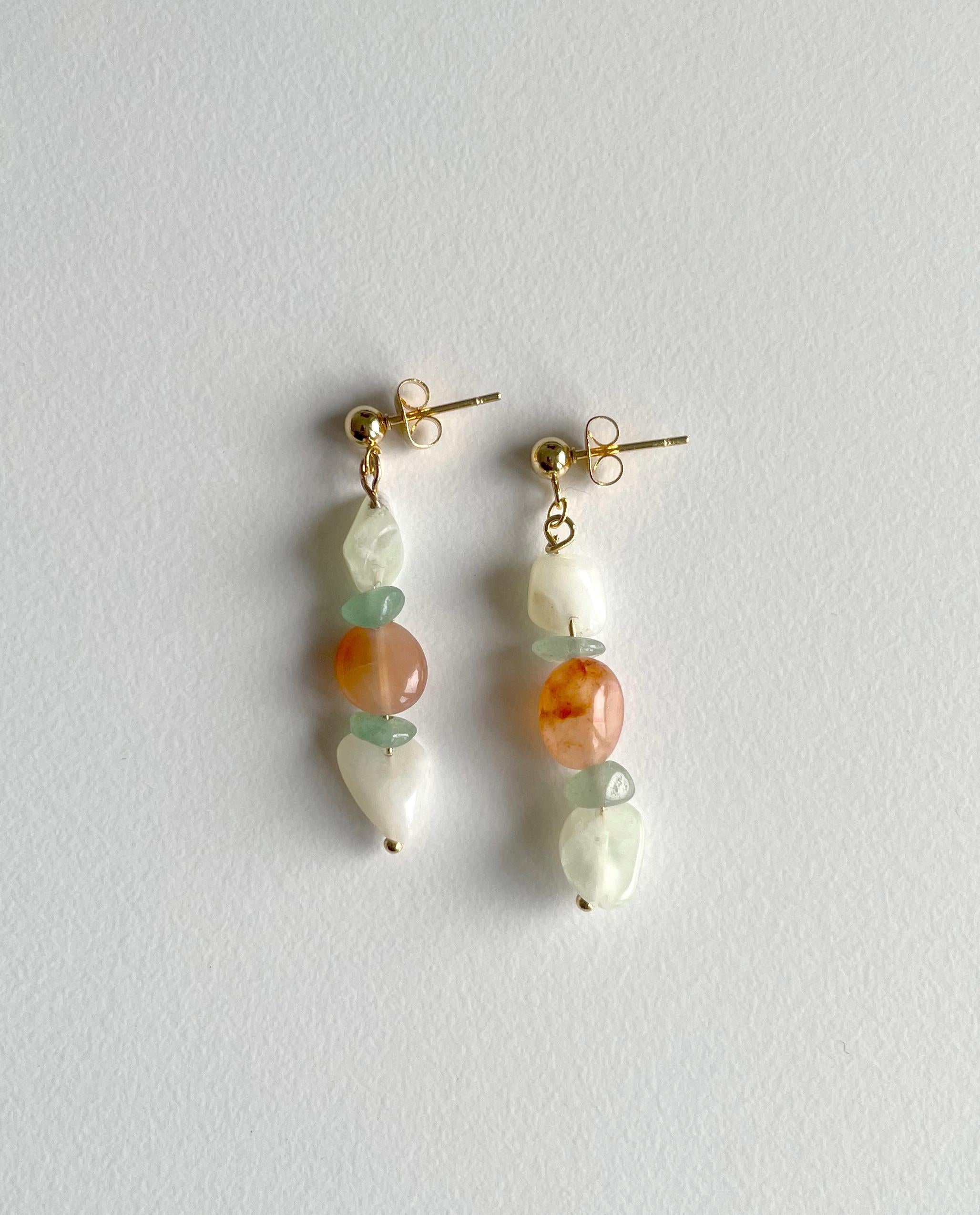 A pair of gold-tone dangle earrings with irregular white beads, light green beads, and round orange stone beads, displayed on a textured light gray background.