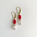 A pair of golden dangle earrings featuring a vibrant red oval bead, small clear natural quartz chip beads, and pale pink round beads, displayed on a clean white textured background.