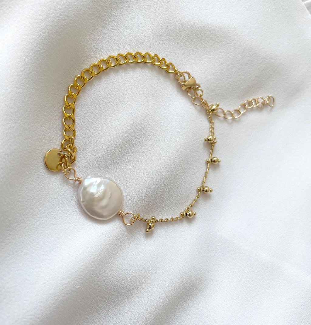 A delicate gold chain bracelet featuring a large, irregular white freshwater pearl and small gold bead accents, displayed on a soft white fabric background.
