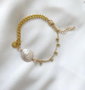 A delicate gold chain bracelet featuring a large, irregular white freshwater pearl and small gold bead accents, displayed on a soft white fabric background.