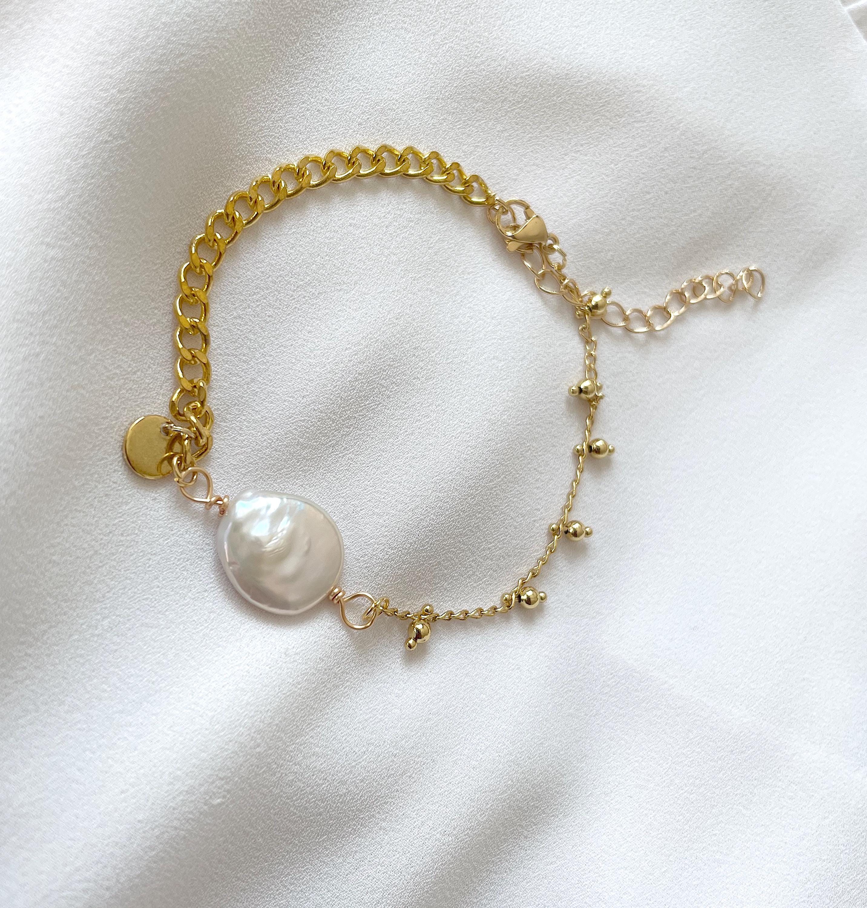 A delicate gold chain bracelet featuring a large, irregular white freshwater pearl and small gold bead accents, displayed on a soft white fabric background.