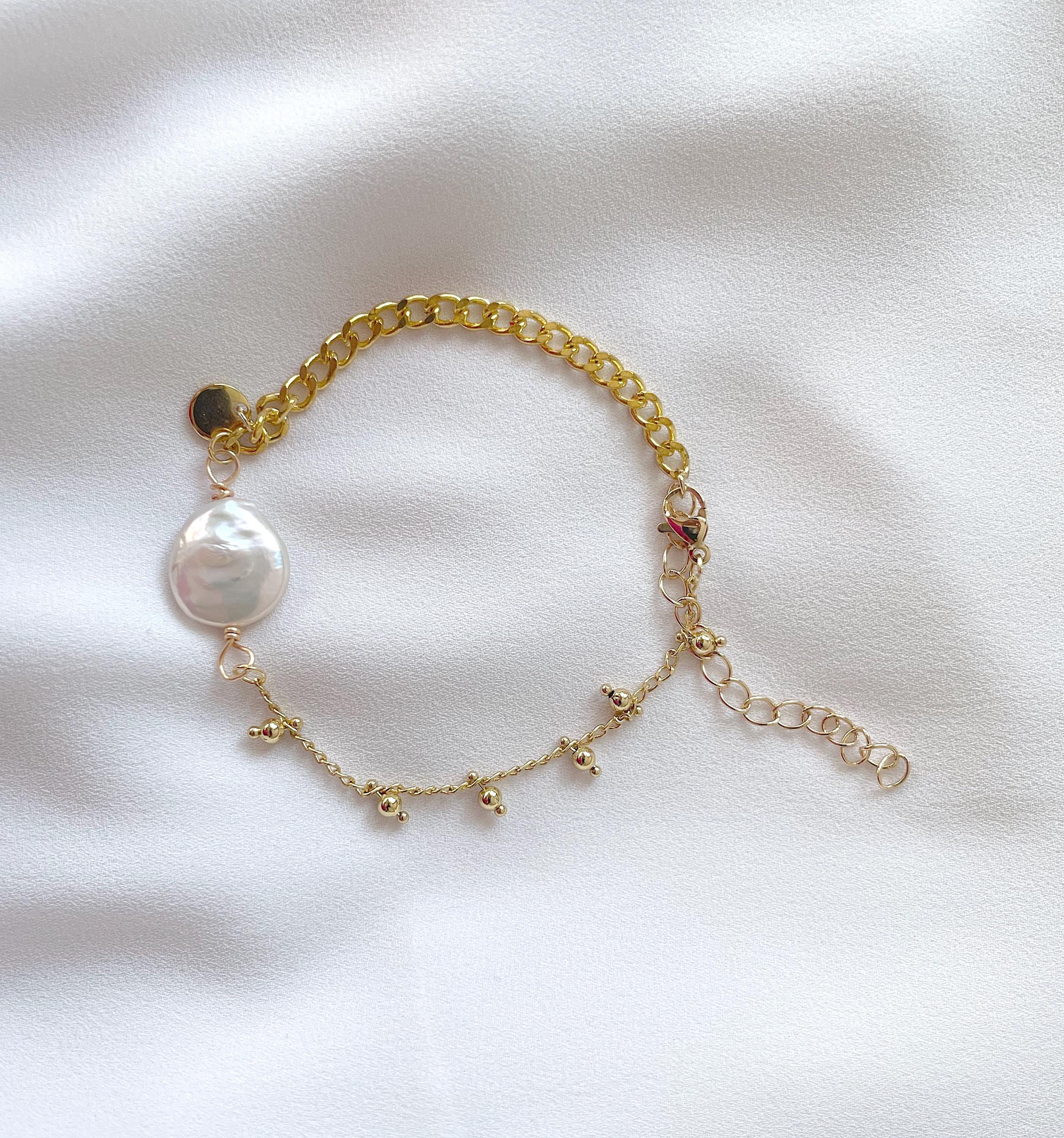 A delicate gold chain bracelet featuring a large white freshwater pearl and small gold star charms, laid flat on a white fabric background.