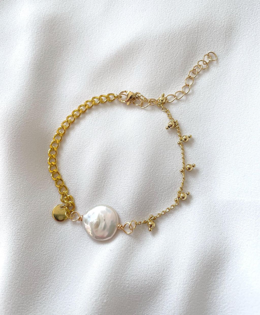 A delicate gold chain bracelet featuring a large, irregular white baroque pearl and small gold charms, laid flat on a white textured fabric background.