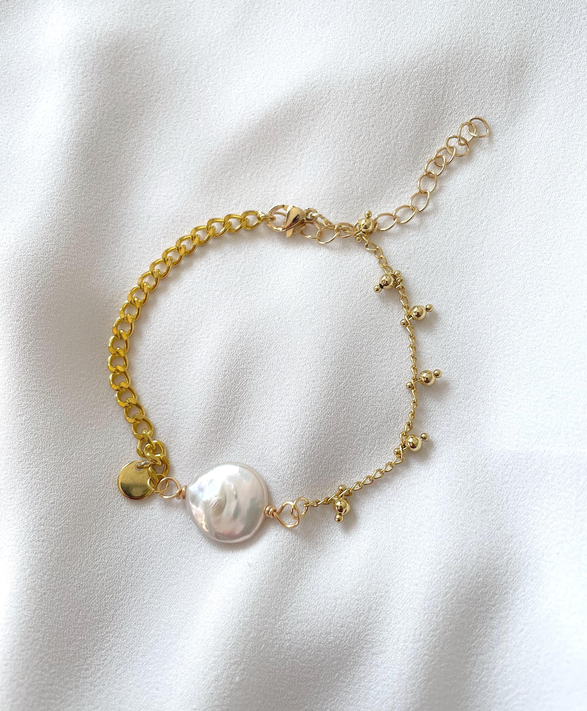 A delicate gold chain bracelet featuring a large, irregular white baroque pearl and small gold charms, laid flat on a white textured fabric background.