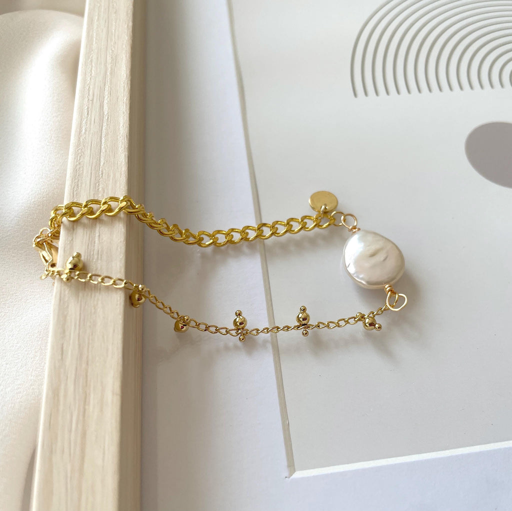 A delicate gold chain bracelet featuring a large, irregular white freshwater pearl and small gold charms, styled on a white surface with a wooden block.