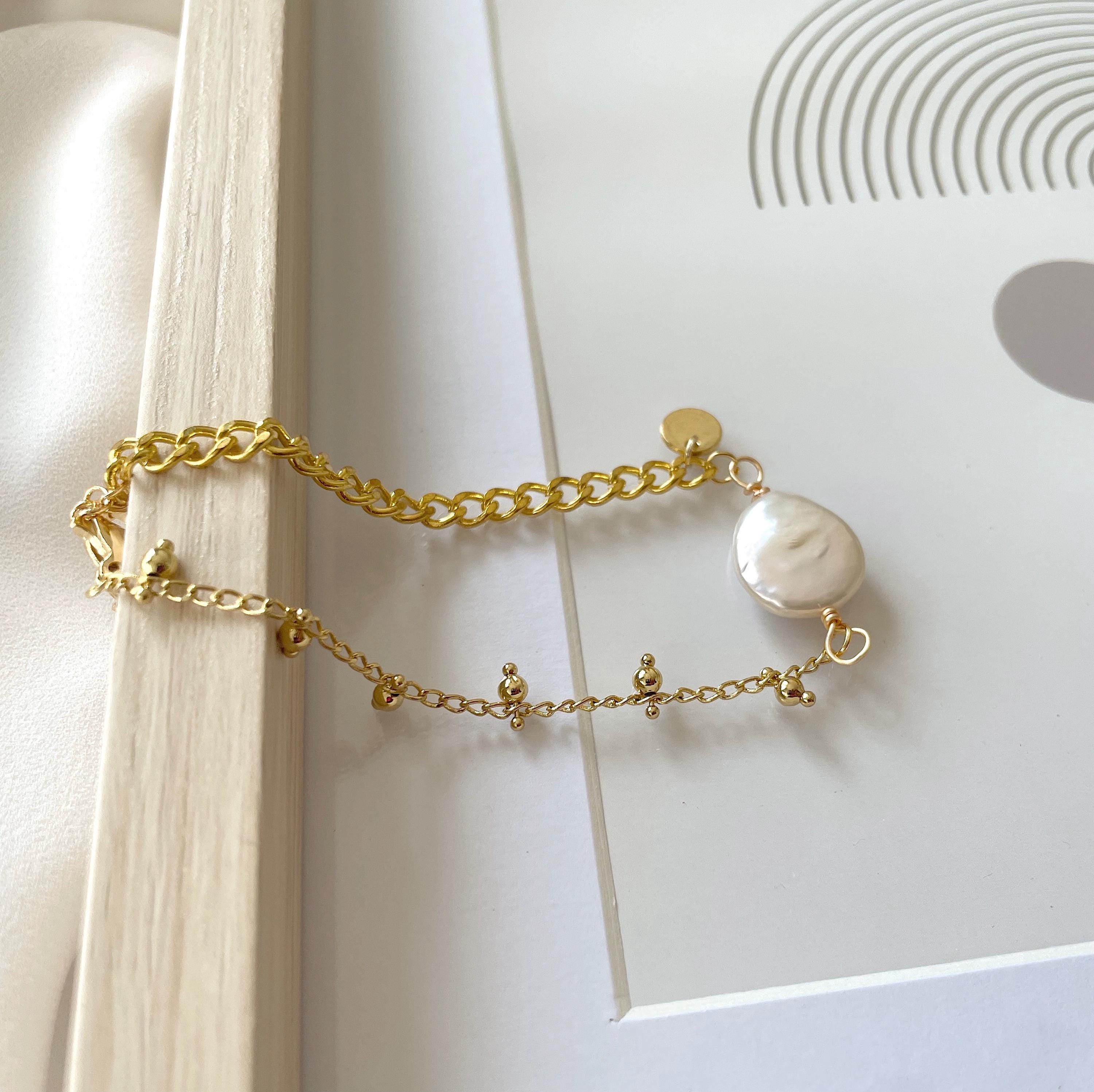A delicate gold chain bracelet featuring a large, irregular white freshwater pearl and small gold charms, styled on a white surface with a wooden block.